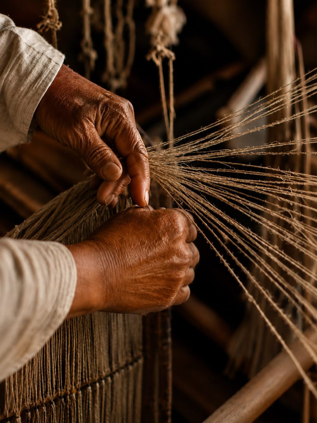 Two weathered hands drawing natural fibre threads onto a hand loom.